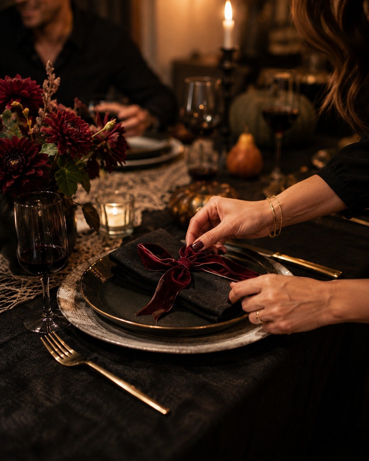 Close-up Halloween place setting with dark plates, a burgundy velvet ribbon, gold flatware, dark flowers, and candlelight.